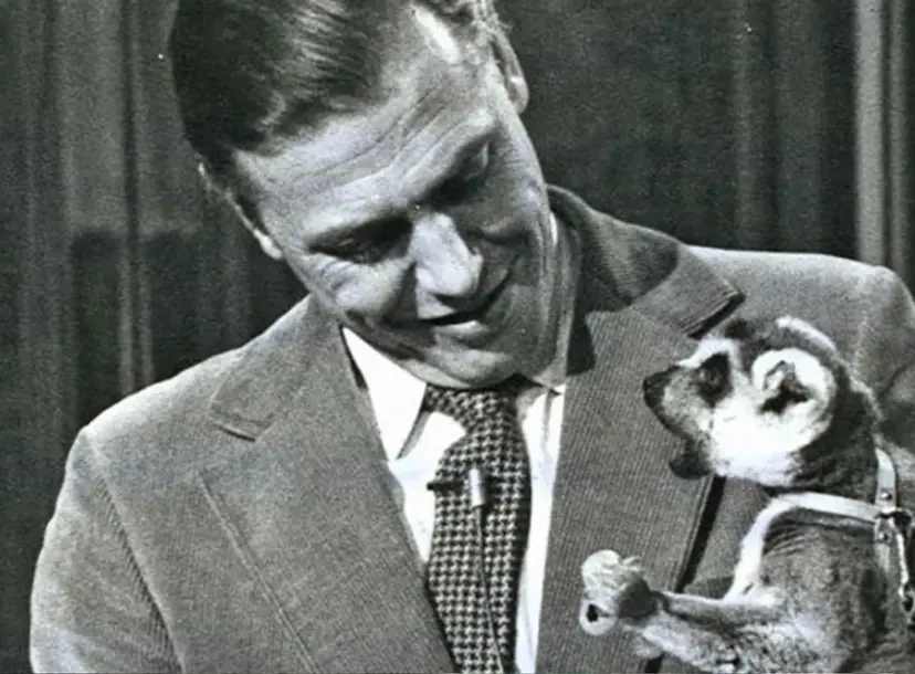 David Attenborough looking down at a lemur that he is holding  