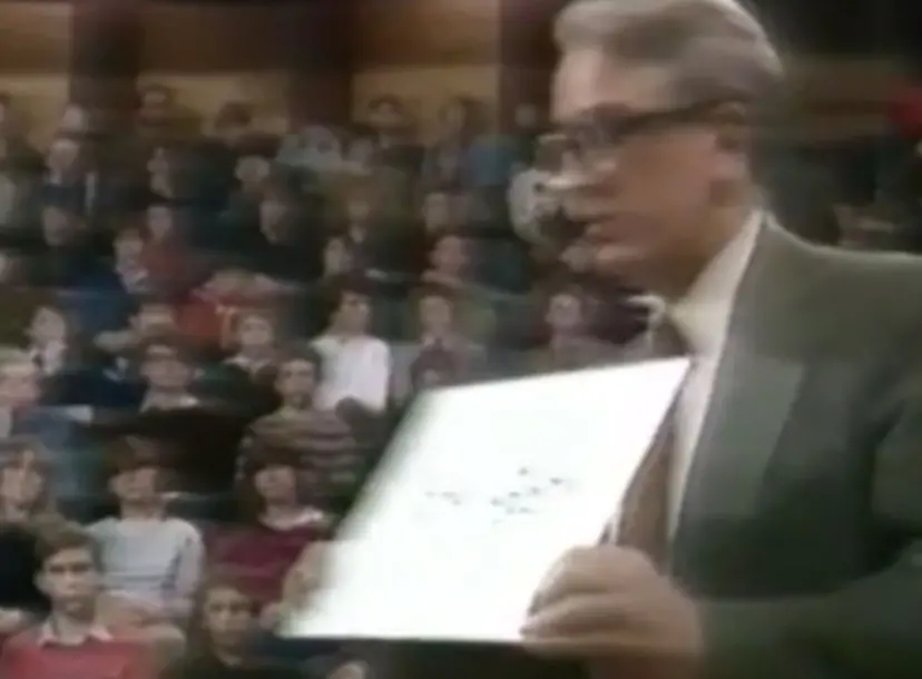 David Phillips holding a sheet of paper in the Ri theatre, a crowd of children watching from their seats