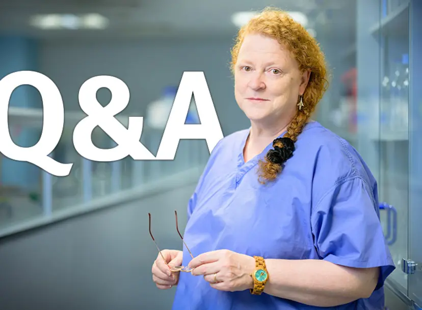 Sue Black standing in a lab, wearing blue scrubs and holding her glasses