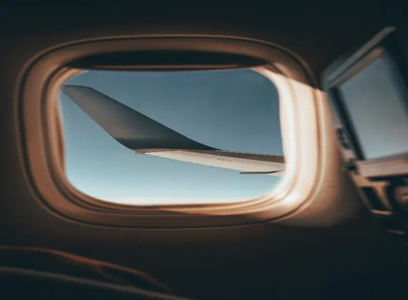 An airplane wing viewed through an airplane window mid-flight, with blue sky in the background.