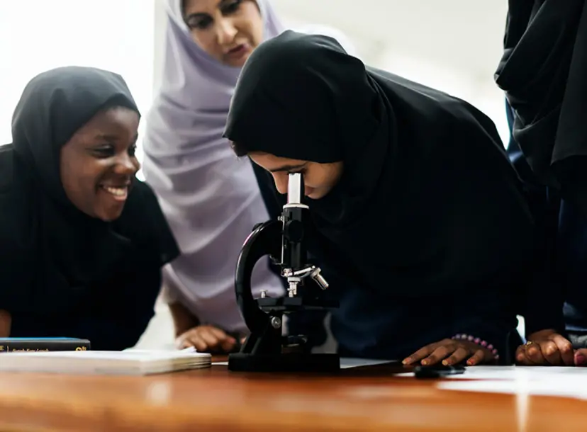 A group of hijabi girls smiles as one looks into a microscope