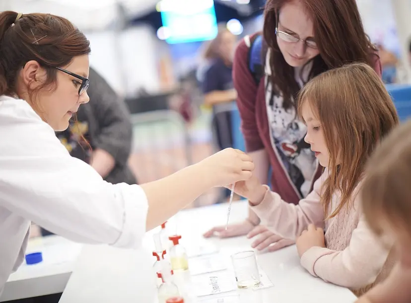 Young people work together with a female scientist dressed in a lab coat, looking at science equipment
