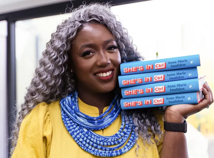 A woman is holding copies of the book 'She's in CTRL' up to her face. She is smiling and wearing a yellow top. 