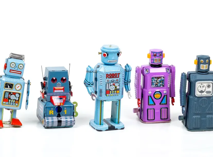Five toy robots are stood in a line. They are all different shapes and colours - light blue, grey, purple and dark blue