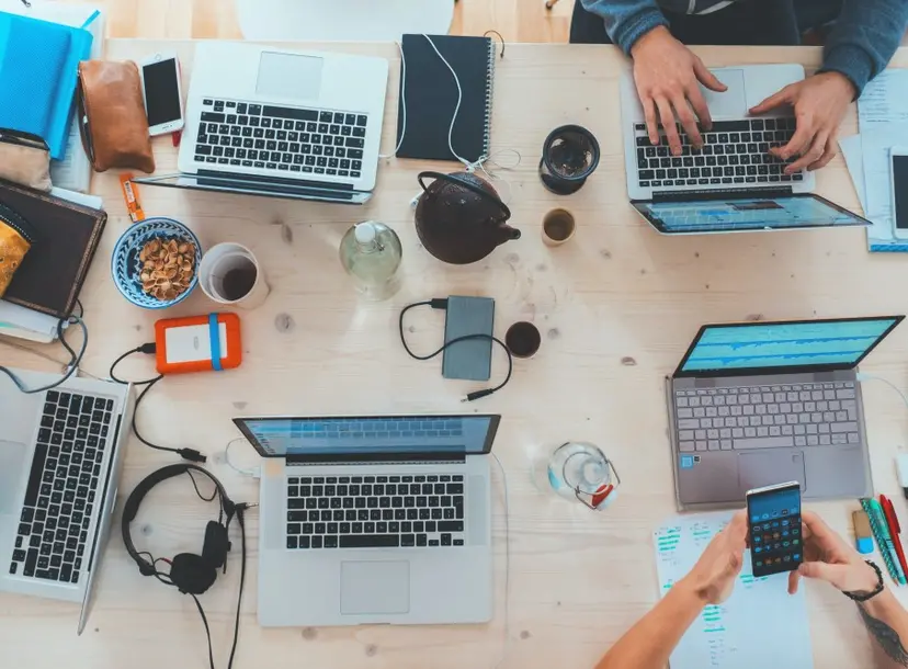 A table top is filled with laptops, chargers, phones and cups of tea and coffee.