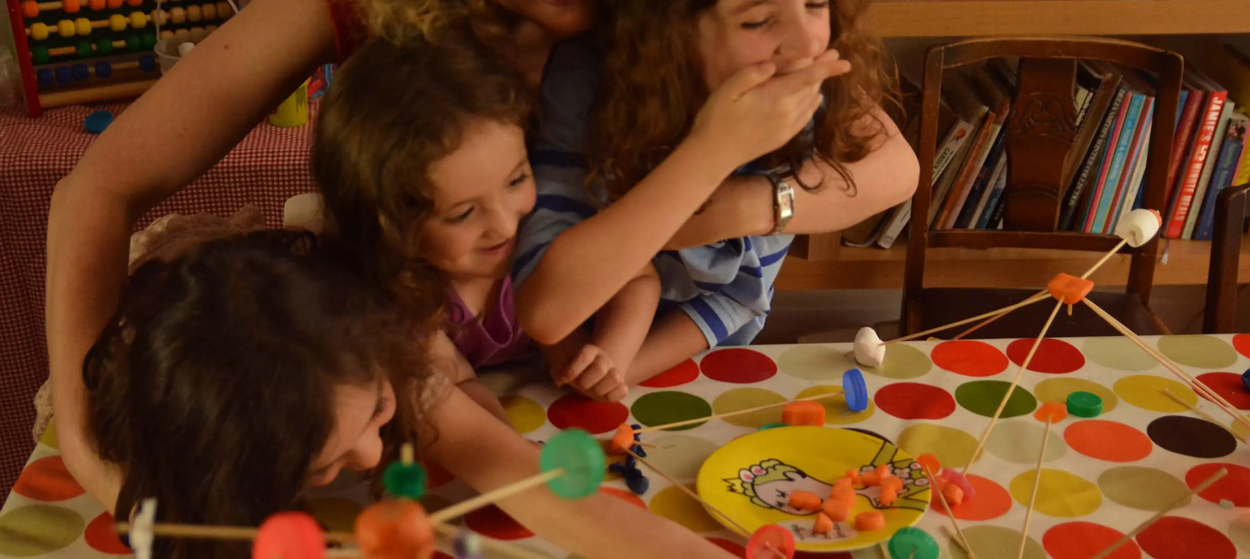three young girls at a kitchen table building a sculpture out of wooden skewers, pieces of carrot and marshmallows