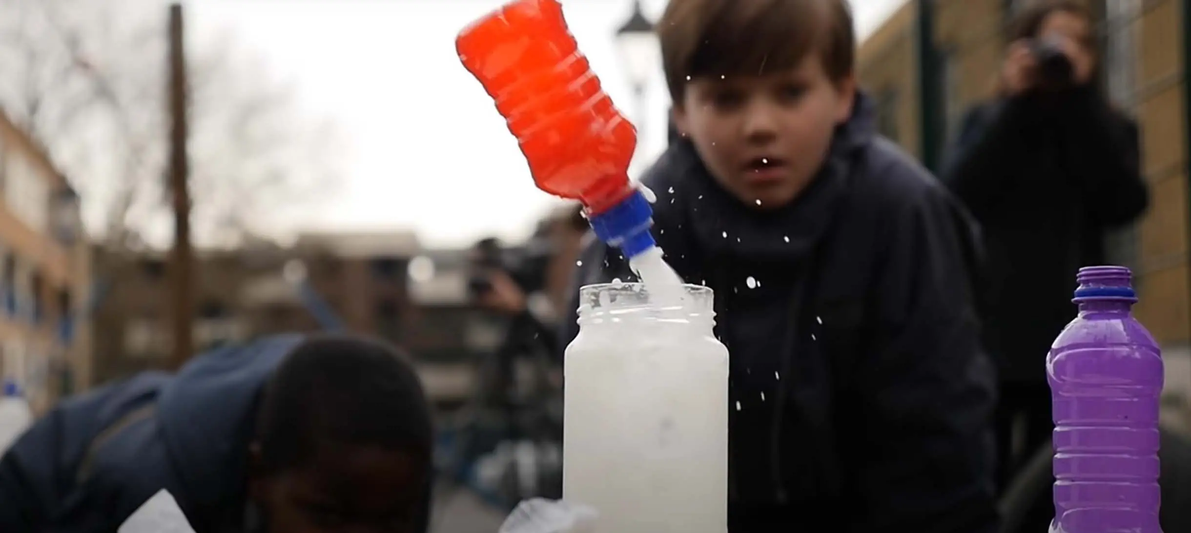 a small fruit juice bottle is being propelled out of a glass jar by a white jet of liquid as a young boy looks on