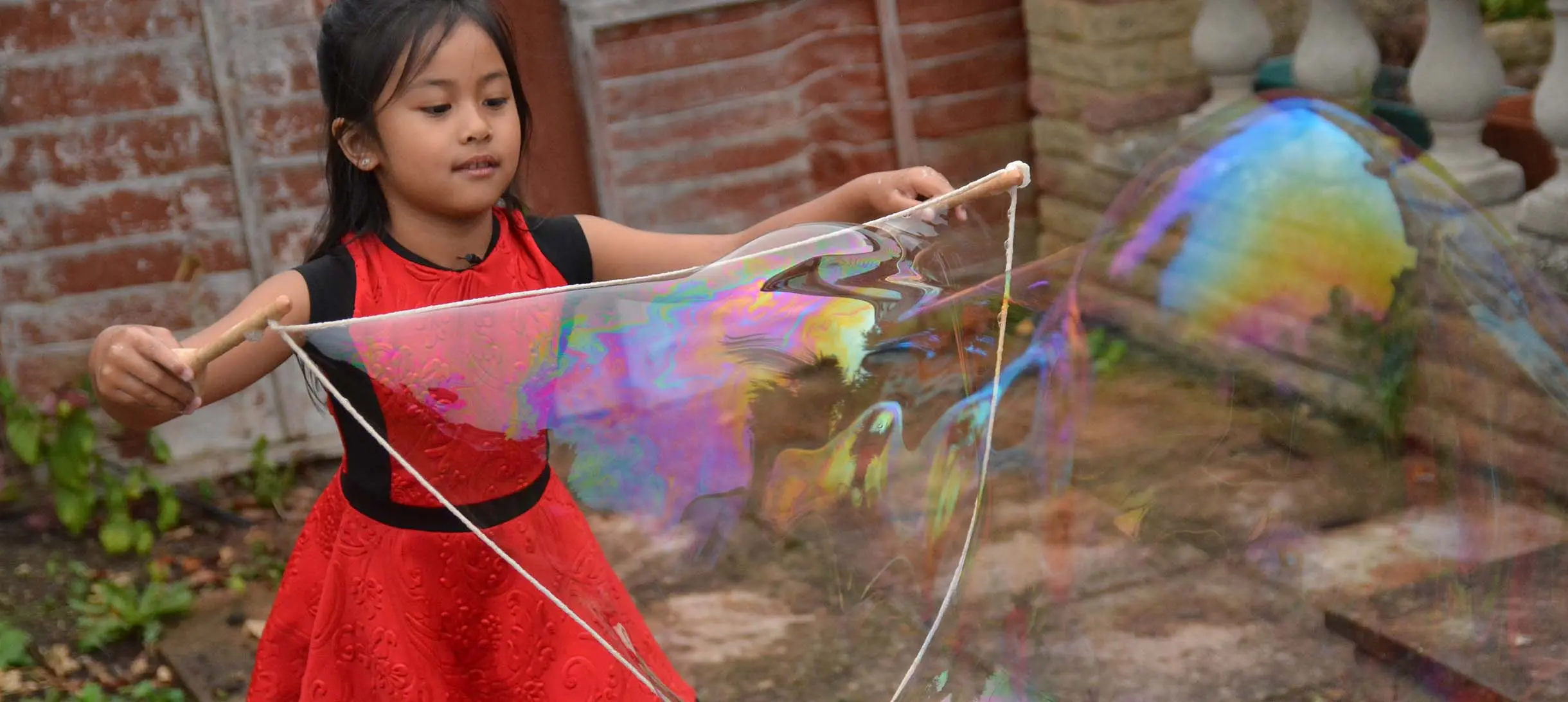 young girl in red dress holding two wooden spoons and some string tied in a loose circle around them from which emerges a giant bubble