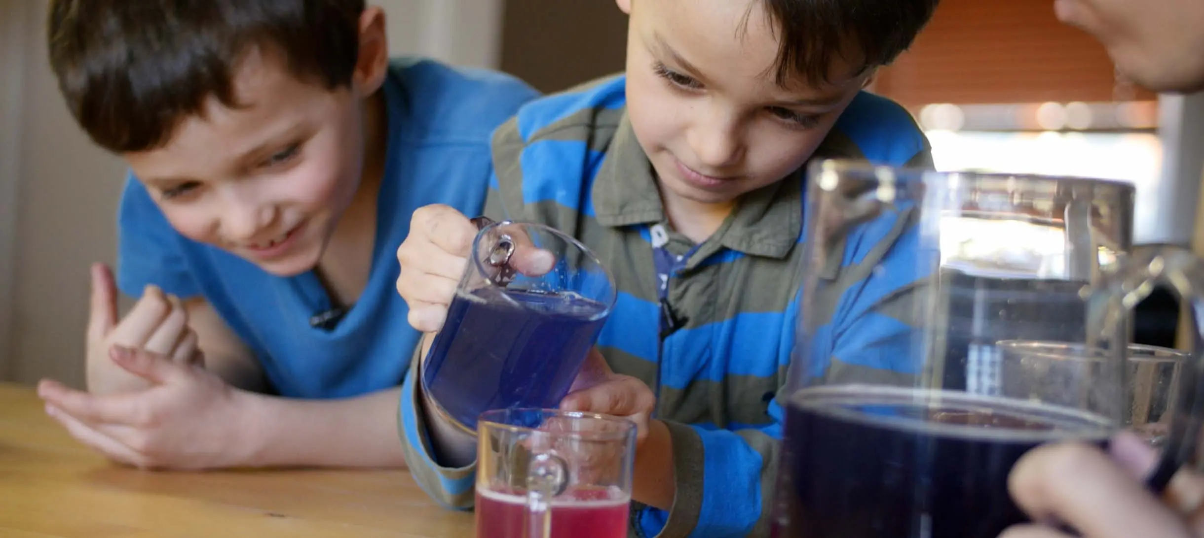 one young boy is pouring a purple liquid into a pink liquid while another boy looks on