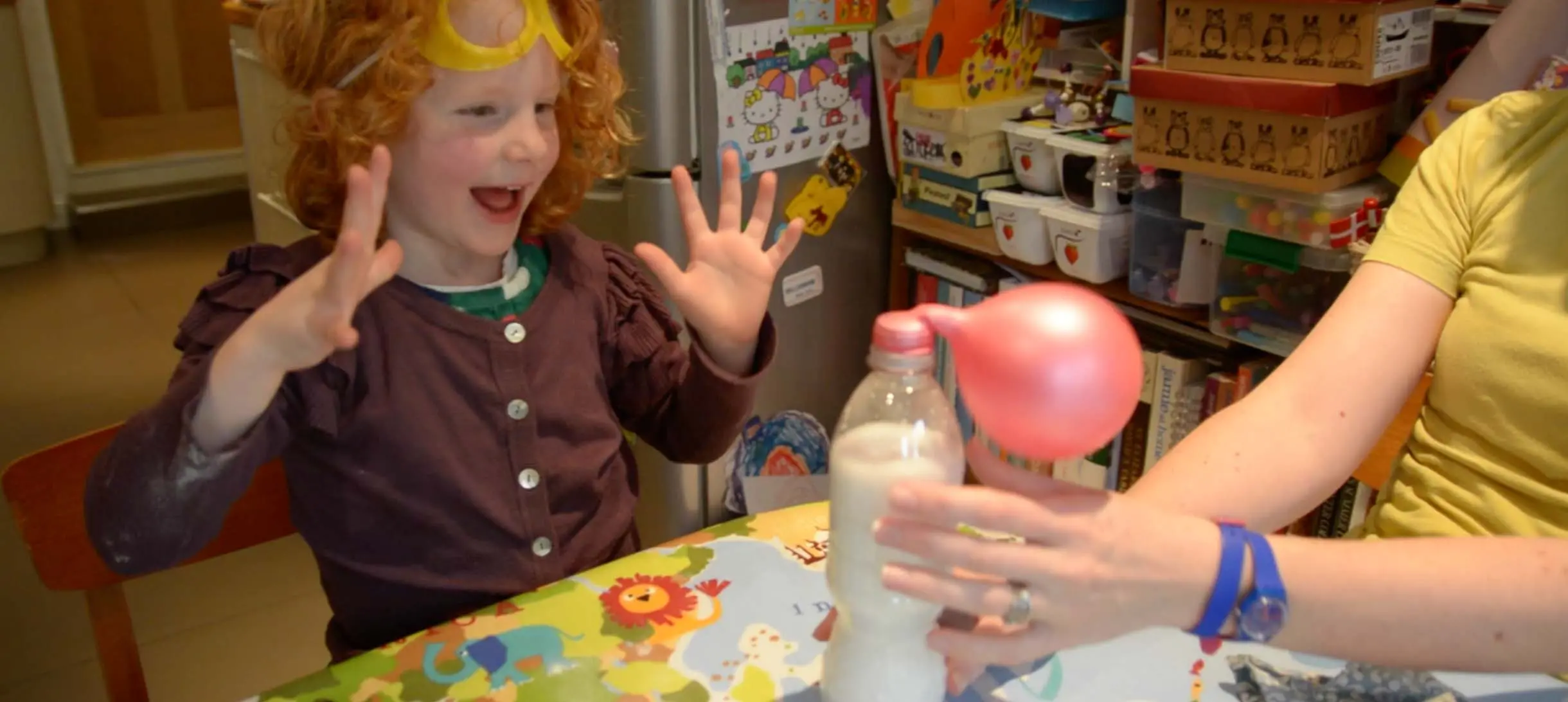 a young girl with curly red hair has her hands up in delighted surprise as she looks at a balloon self-inflating