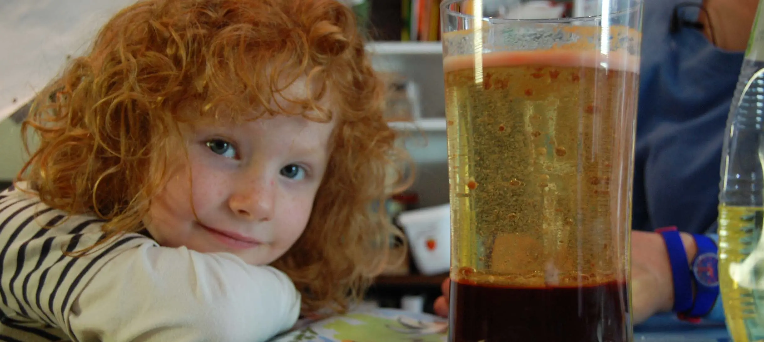 red curly haired young girl smiles to the camera, next to her is a glass filled with oil and some red liquid