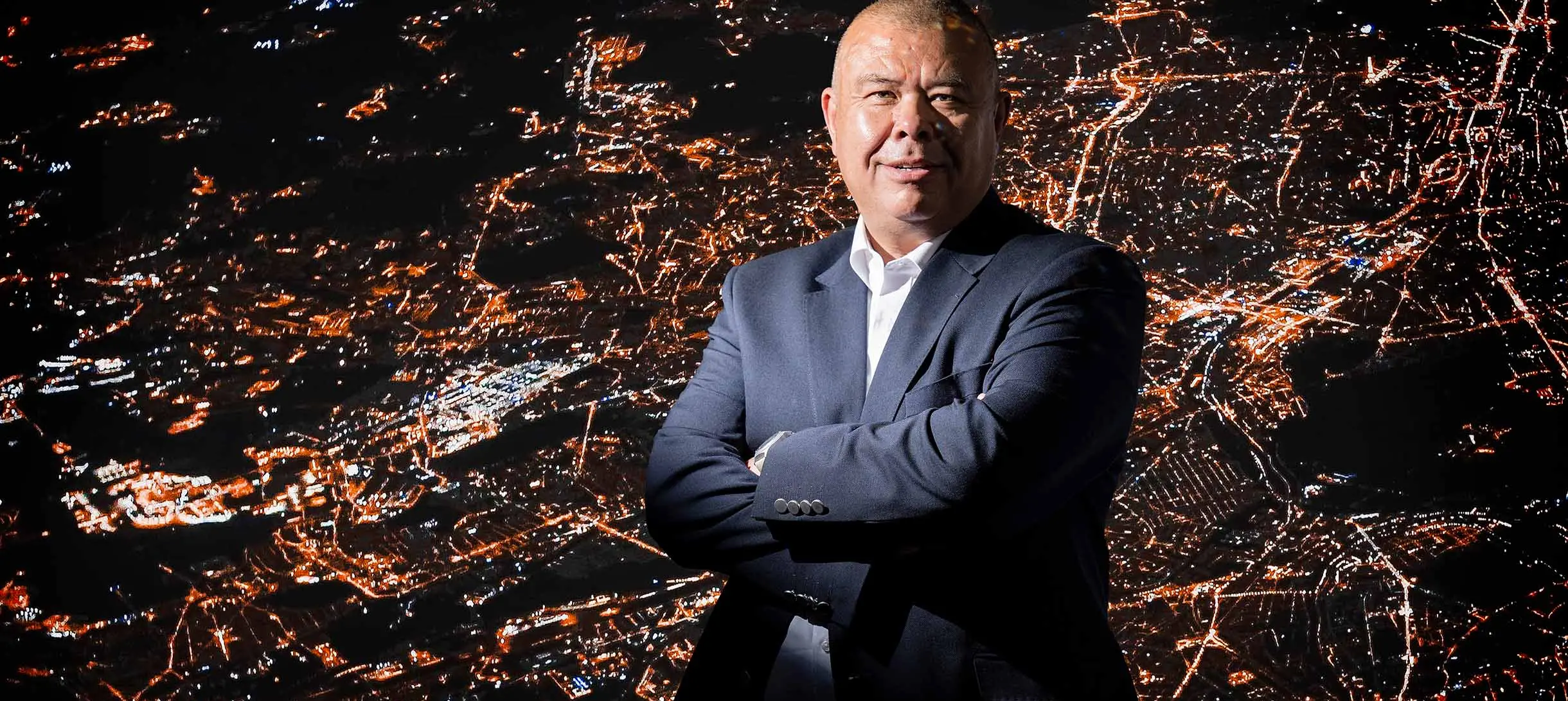 Jonathan Van-Tam with his arms crossed in front of an aerial view of a city at night with bright lights