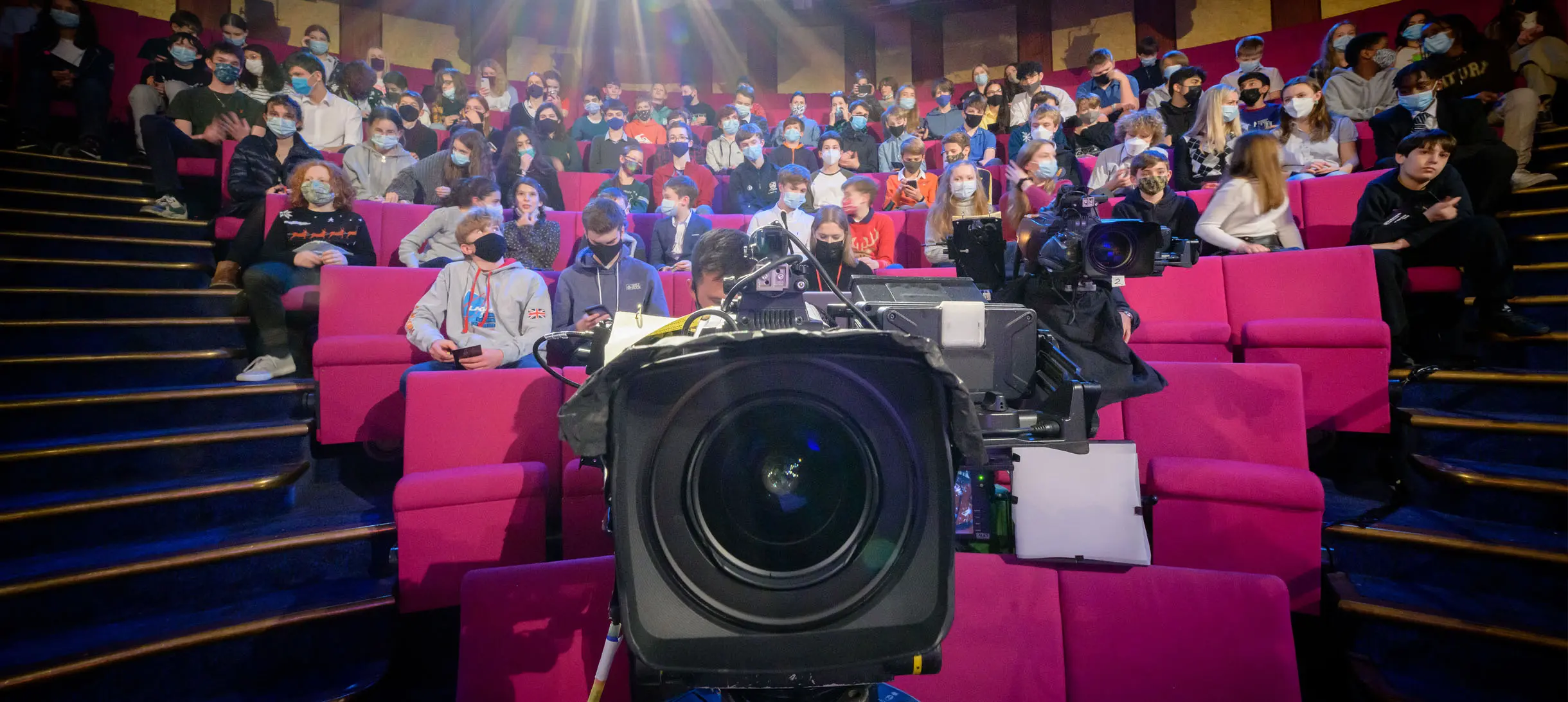 Image of a film camera lens with an audience sitting behind in a Theatre