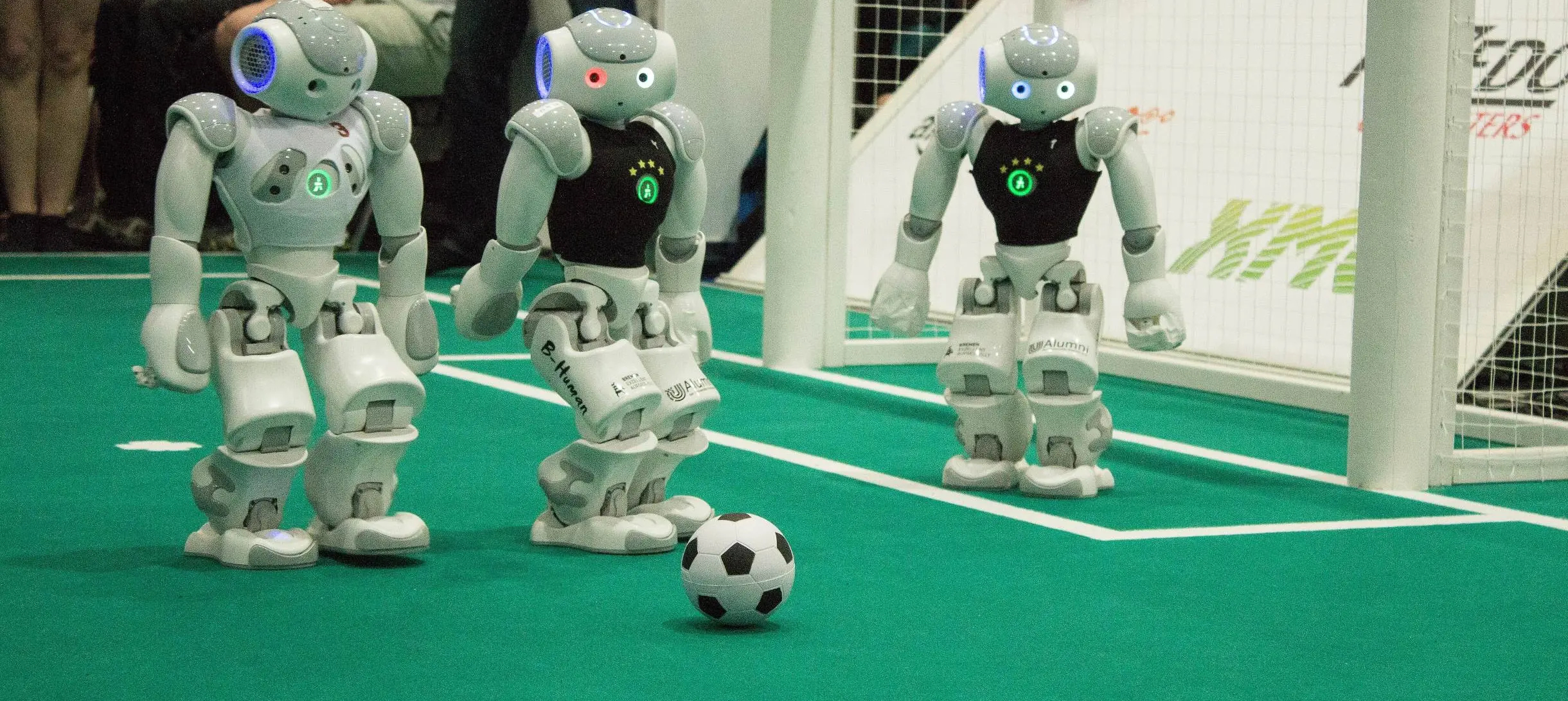 Three robots stand behind a football on an artificial football pitch, one of them is stood inside a goal.