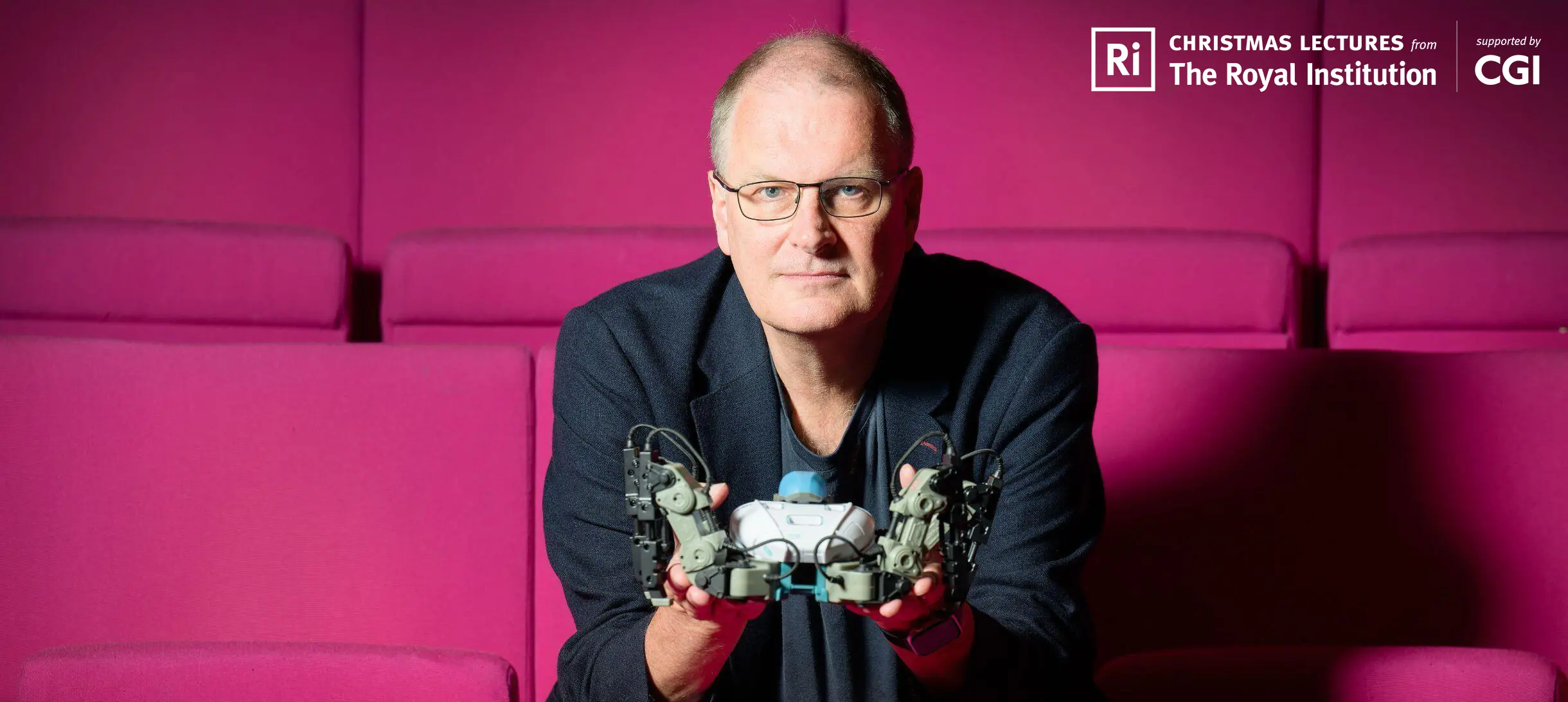 Mike looks into the camera while holding a small robot, sitting on red seats in our Theatre