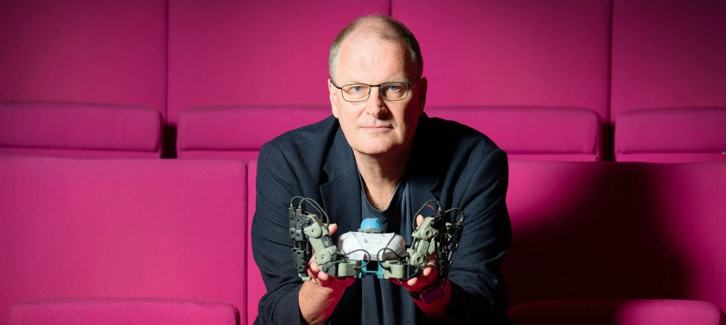 Mike looks into the camera while holding a small robot, sitting on red seats in our Theatre