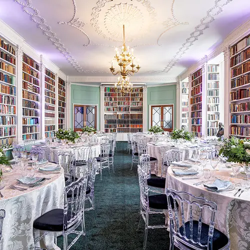 a large room with bookshelves lining all the walls, in the middle of the room are six round tables with flower arrangements on them and chairs around