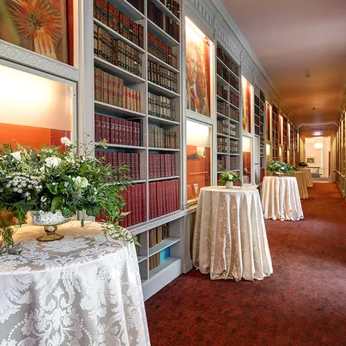 a long corridor with red carpet and bookshelves lining the walls, there are small round tables dotted along the length of the corridor