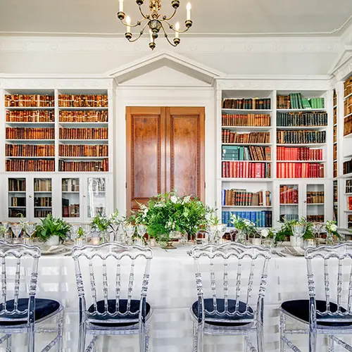 A small room with bookshelves lining the walls and a long dining table in the middle with chairs on either side. The table is covered in a white table cloth and there are flower arrangements on the table.