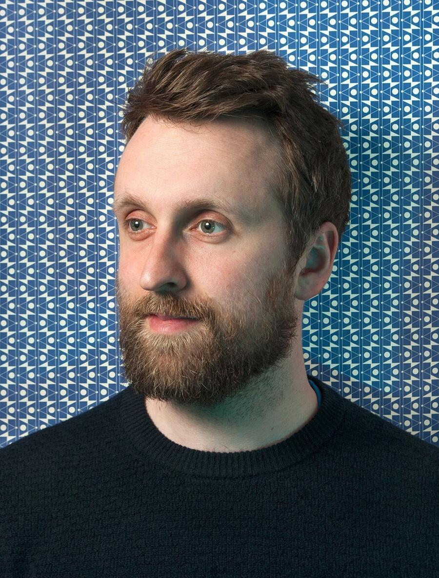 Side profile of a man with brown hair and a beard, wearing a black top.