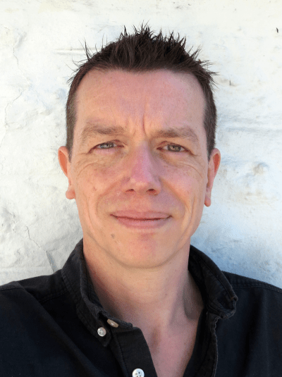 A man slightly smiling with short dark hair wearing an unbuttoned black shirt