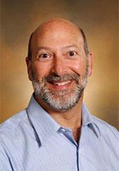 David is smiling towards the camera. He is wearing a pale blue shirt.