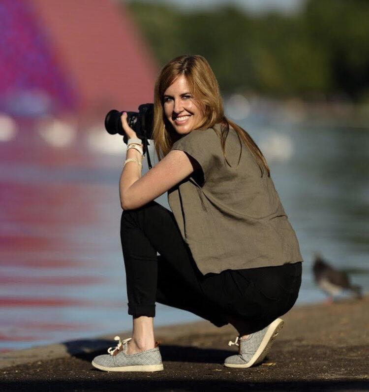 A woman is bending down on her knee with a camera in her hand. She is smiling and facing the screen.