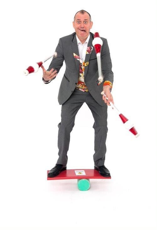 A white man is balancing on a board, that is ontop of a roller. He is also juggling at the same time.