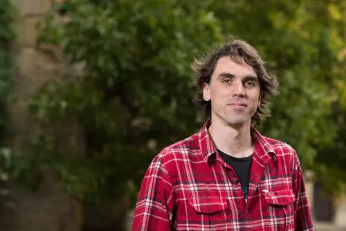 A photo of a white man with shoulder length dark hair wearing a red checked shirt