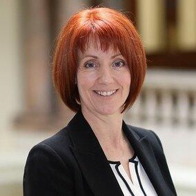 A woman with short red hair is looking at the camera. She is wearing a black blazer and is smiling.