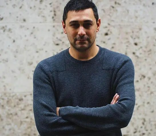 Adam is looking at the camera, wearing a blue jumper. His arms are crossed over his chest.