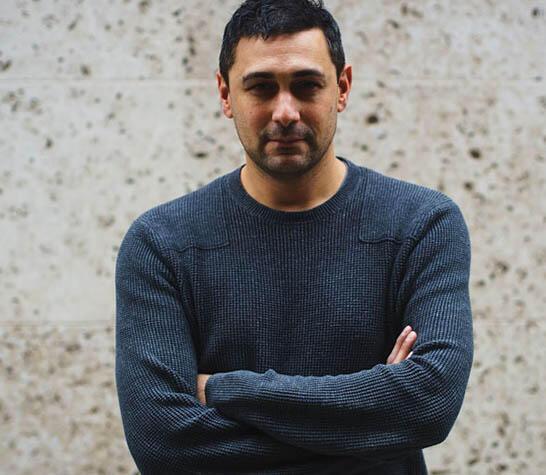 Adam is looking at the camera, wearing a blue jumper. His arms are crossed over his chest.
