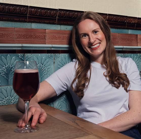 A woman faces the screen, holding a beer.