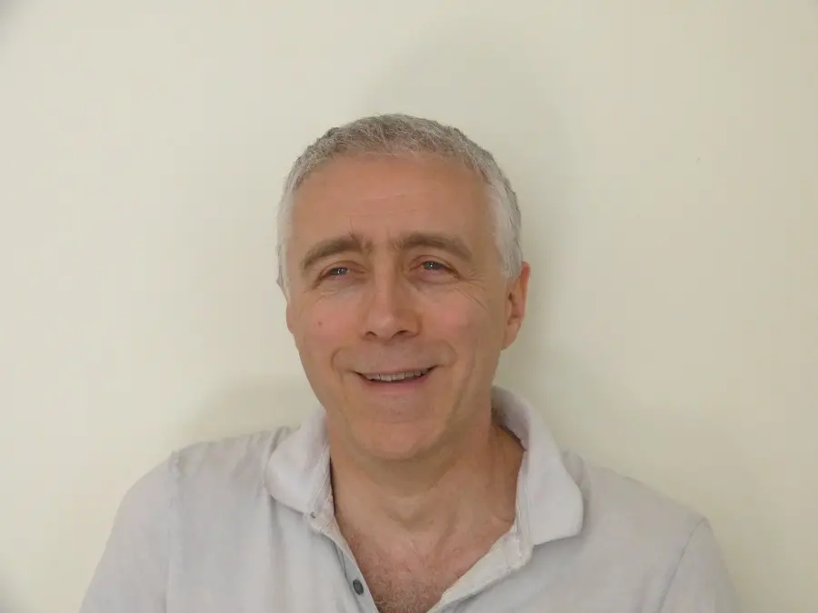 A man with short white hair is standing in front of a white wall. He is wearing a white shirt.