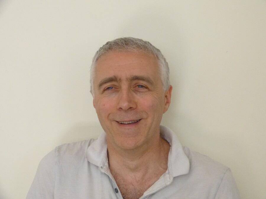 A man with short white hair is standing in front of a white wall. He is wearing a white shirt.