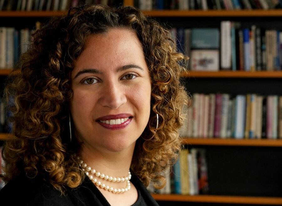A woman with dark curly hair is smiling towards the camera. She is wearing a white necklace.