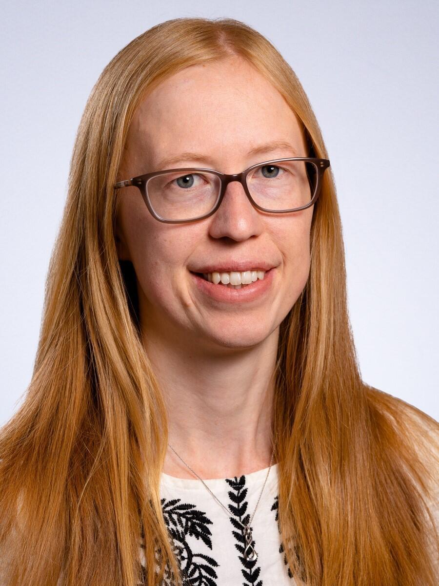 A woman with copper coloured hair is smiling towards the camera. She is wearing dark glasses.