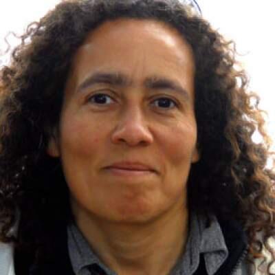 A woman with dark curly hair is looking rowards the camera. She is smiling.
