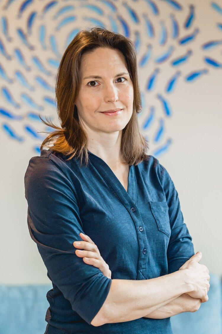 A woman with brown hair is looking towards the camera. She is wearing a blue top and he arms are crossed across her body.