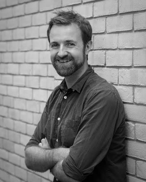 A black and white photo of a man stadning against a wall. He is smiling.