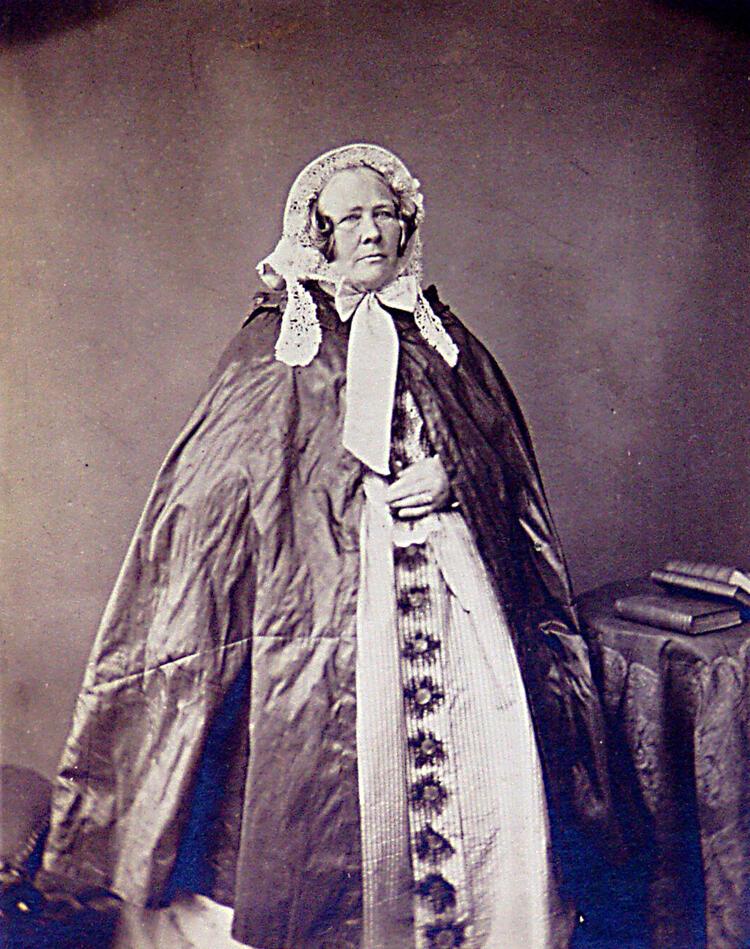 A black and white photo of Anne Phillips, wearing a big white dress, a dark cloak and a white bonnet