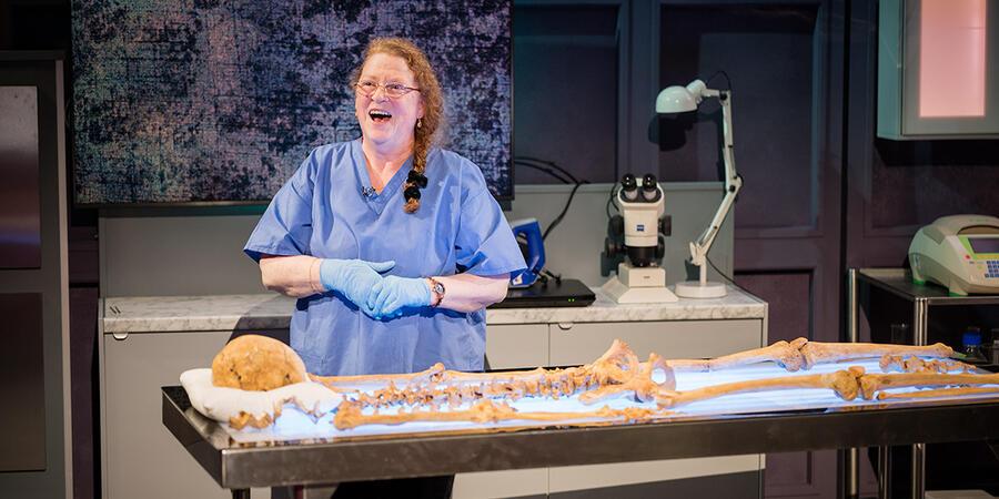 Sue Black is standing behind a table with a skeleton laid out on it, smiling. She is wearing blue scrubs and gloves.
