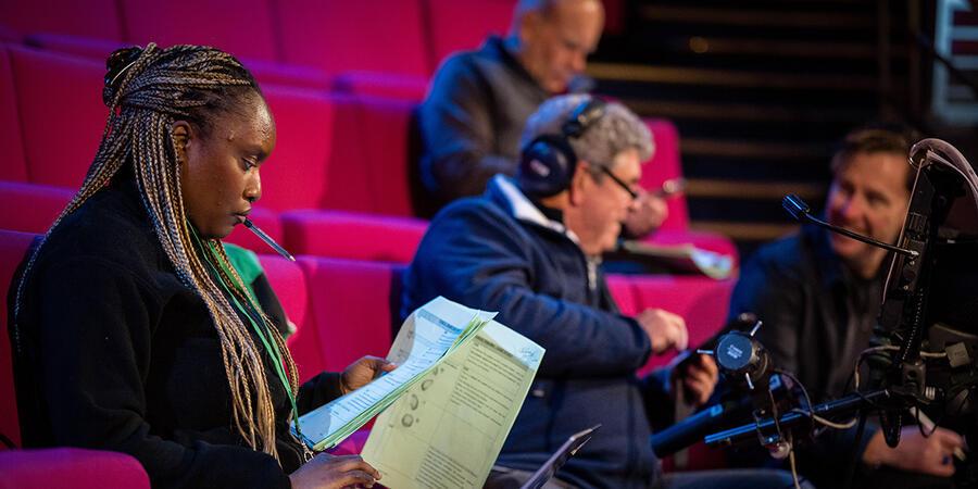 Production crew sitting on the magenta seats. We can see a woman reviewing a script, and men talking over a large video camera