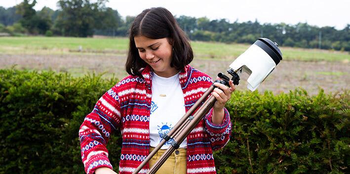 Mia is holding a white cylindrical apparatus on a large tripod