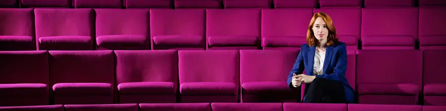 Hannah Fry wearing a blue suit, sitting among the magenta seats in the Ri Theatre