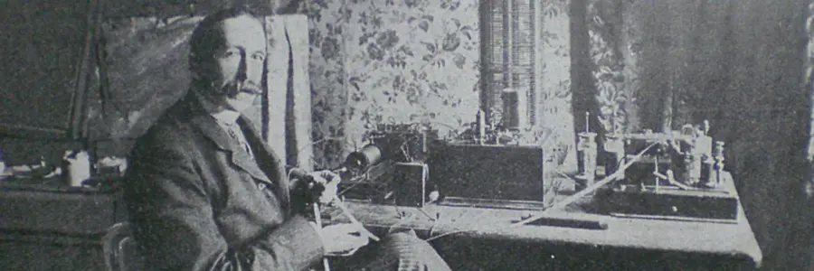 Nevil Maskelyne, sporting a great big moustache, sitting at a table crowded with equipment