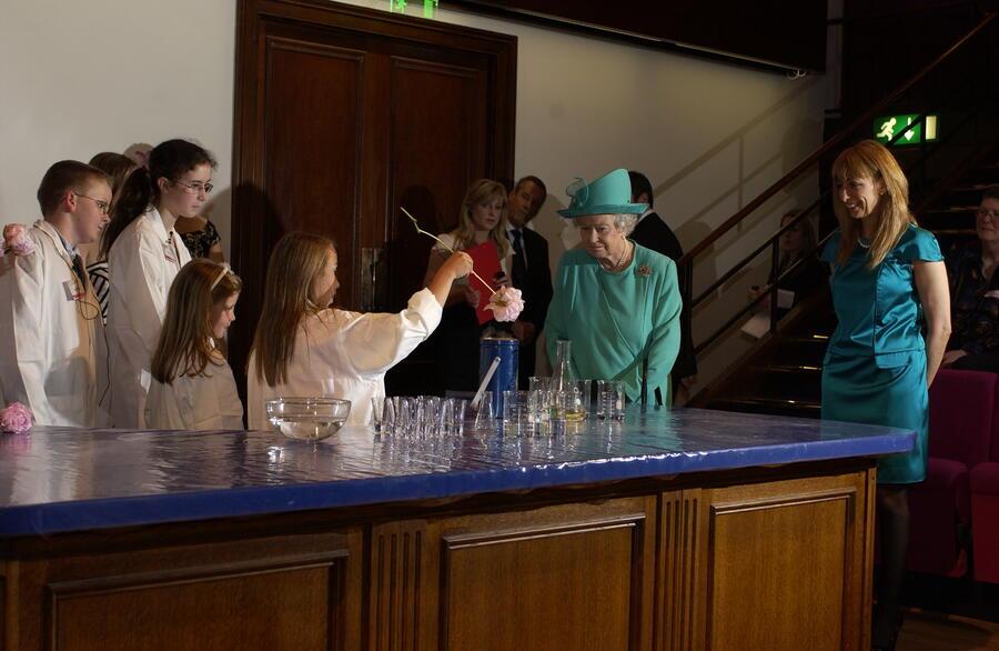 The Queen wearing a green dress and hat, watching a child give a science demonstration with dry ice