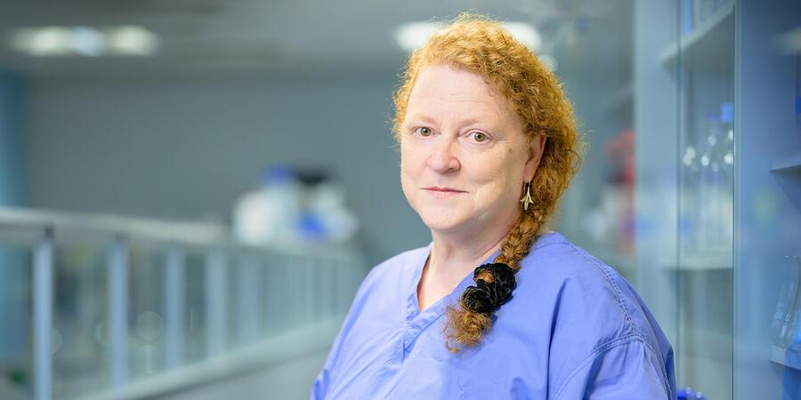 Sue Black standing in a lab, wearing blue scrubs. Her curly red hair is in a plat.