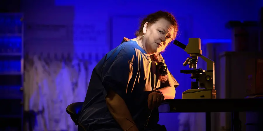 Sue Black wearing scrubs, sitting at a table with a microscope