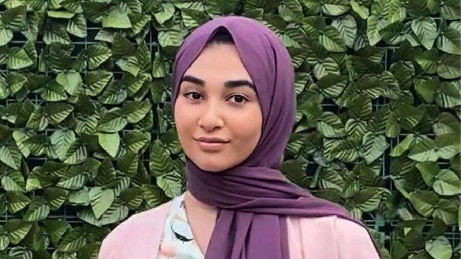 A picture of Zara Hussan posing in front of a leafy hedge. She wears a pink top with a purple hijab and looks into the camera.